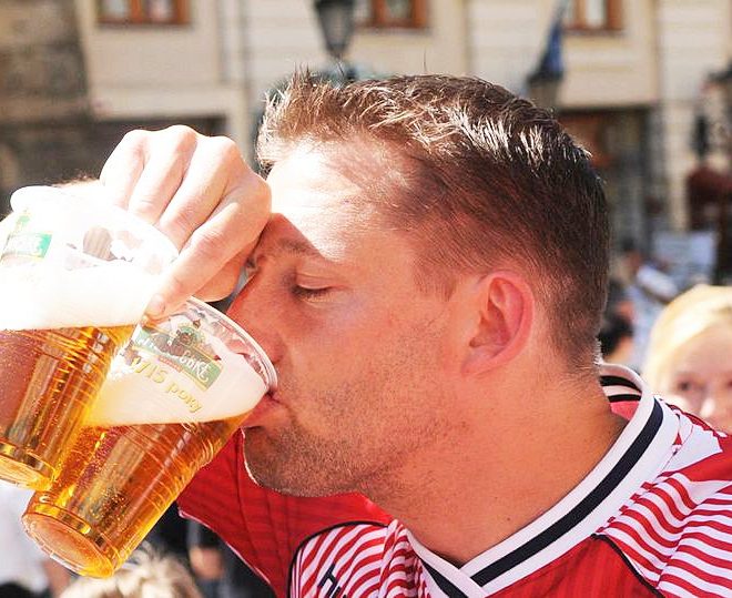 a football fan drinks from one of the two cups of beer he is holding in his right hand